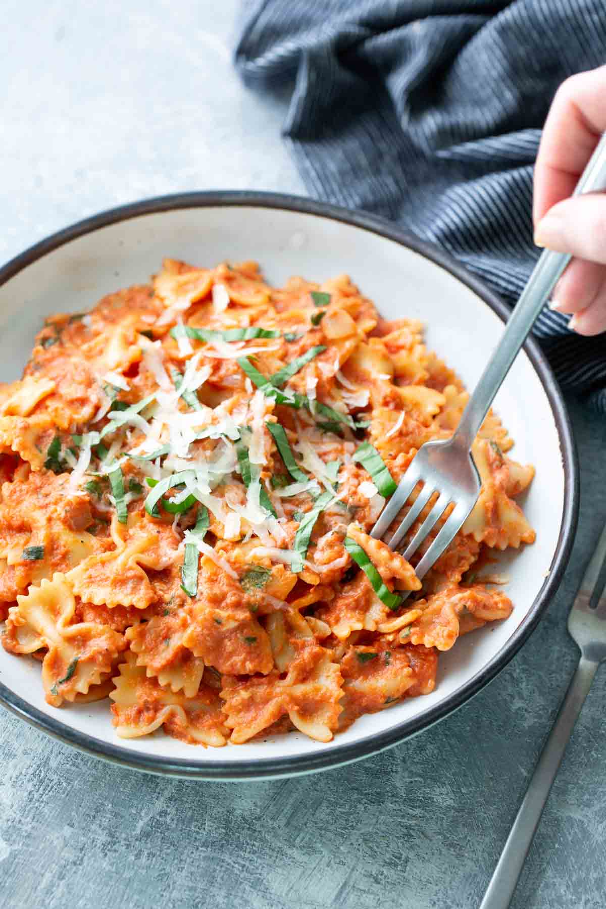A bowl of farfalle pasta with tomato sauce, topped with grated cheese and basil, with a hand holding a fork ready to eat.