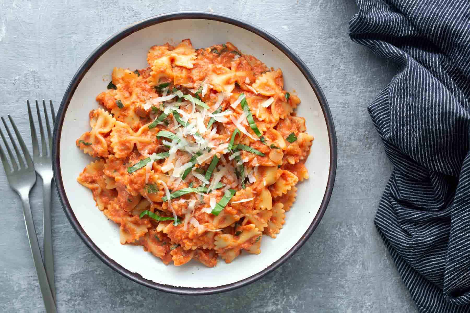 A bowl of bow tie pasta with tomato sauce, garnished with shredded cheese and basil, next to two forks and a dark striped napkin on a gray surface.