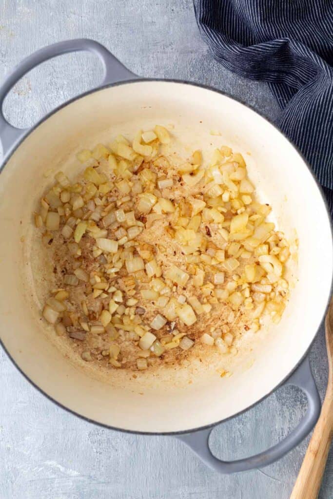 Chopped onions are sautéing and browning in a large white pot, with a wooden spoon and a dark striped cloth nearby.