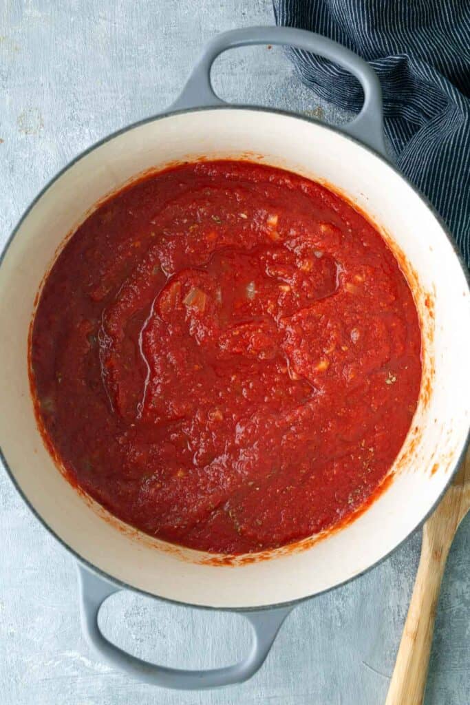 A pot filled with red tomato sauce sits on a grey surface next to a wooden spoon and a dark striped cloth.