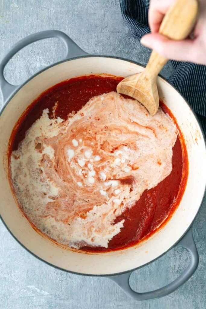 A hand stirs cream into a pot of tomato sauce with a wooden spoon, blending the ingredients together on a gray surface.