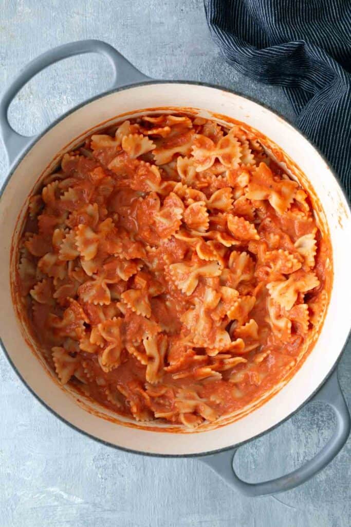 A pot of farfalle pasta mixed with tomato cream sauce, viewed from above, on a light gray surface with a striped cloth nearby.