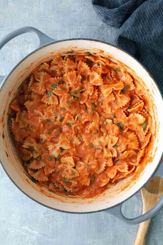A large pot filled with bowtie pasta mixed in a creamy tomato sauce with herbs, next to a wooden spoon and a blue-striped cloth.