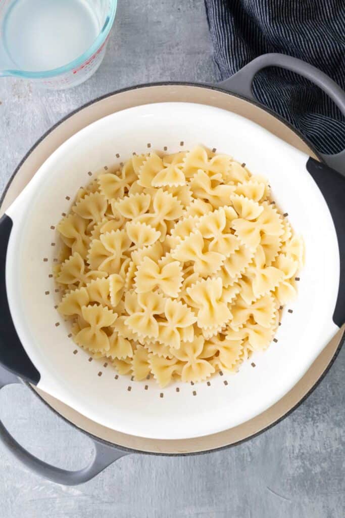 Cooked bow tie pasta is draining in a white colander placed over a pot, with a measuring cup of liquid and a striped cloth nearby.