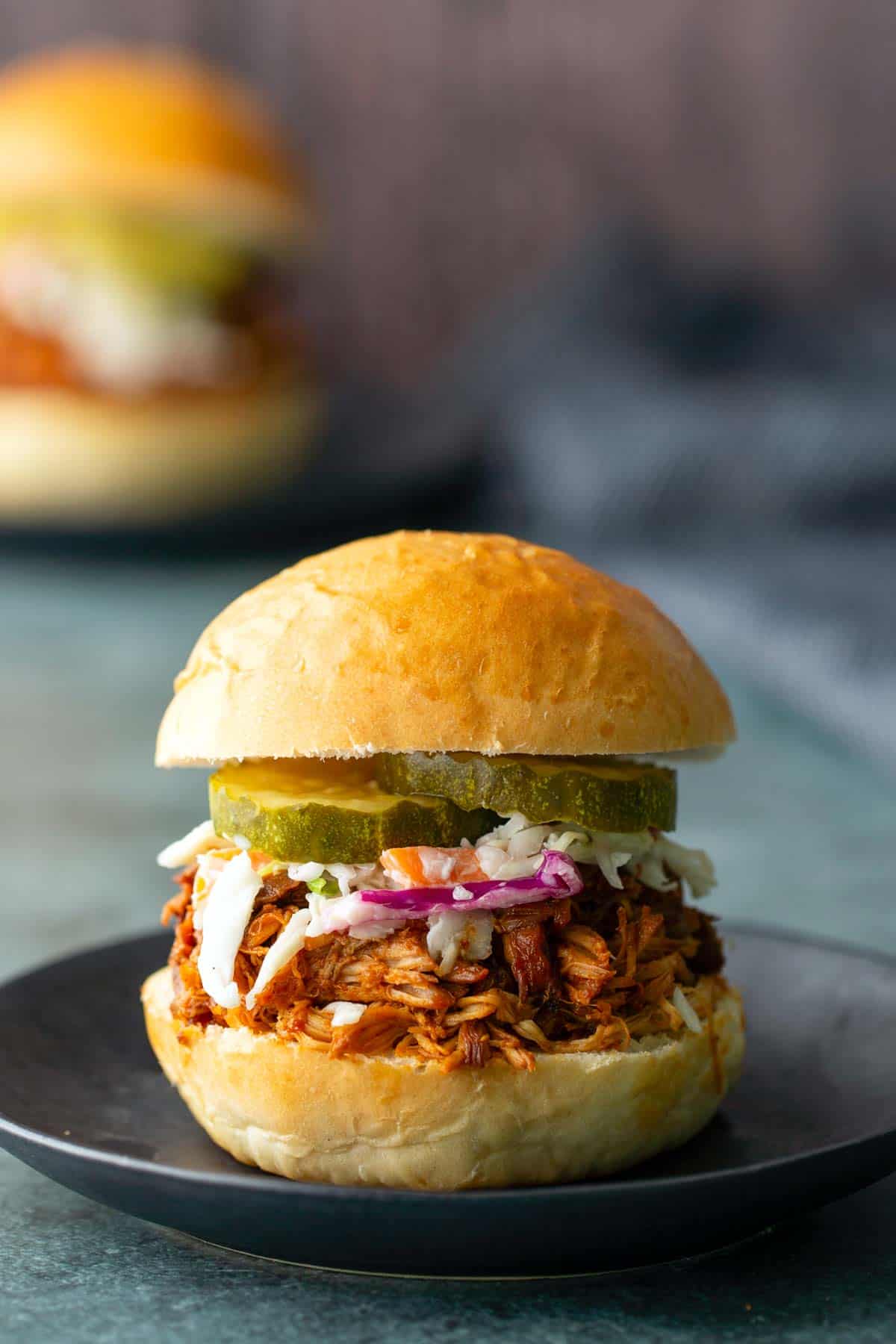 A pulled pork sandwich topped with coleslaw and pickle slices sits on a black plate, with another bbq chicken sandwich blurred in the background.