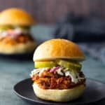 Pulled pork sandwich with coleslaw and pickles on a bun, served on a black plate alongside a bbq chicken sandwich blurred in the background.