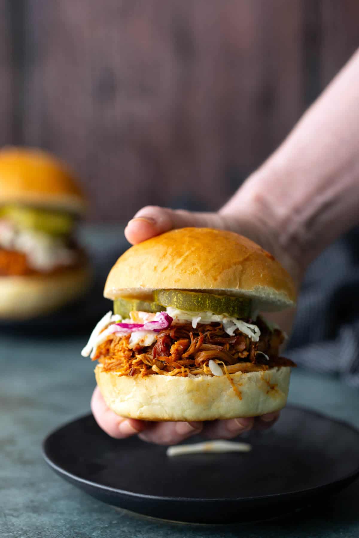 A hand holds a mouthwatering bbq chicken sandwich with coleslaw and pickle slices nestled in a soft bun, while another similar sandwich appears blurred in the background.