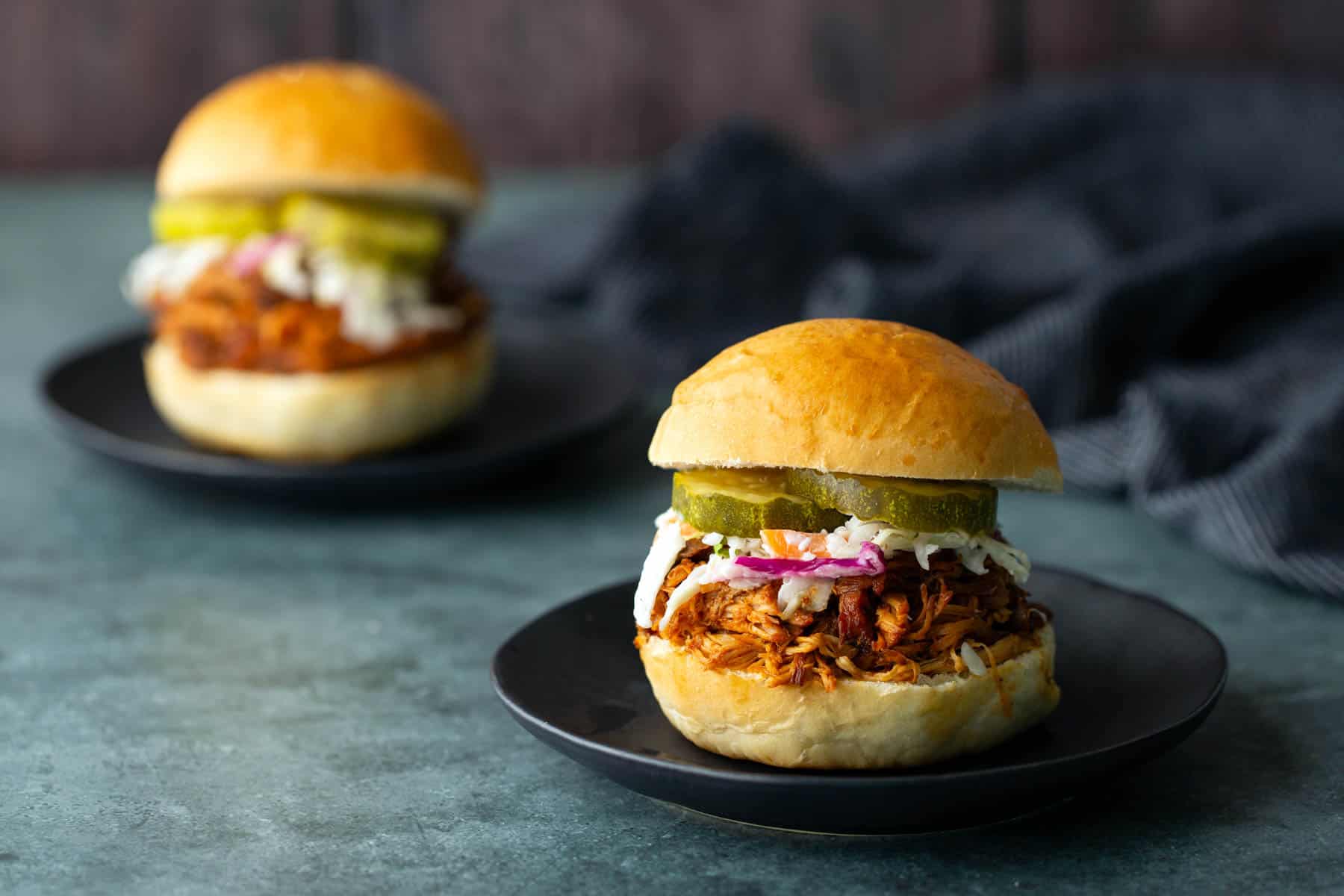Two pulled pork sandwiches on black plates, topped with coleslaw and pickle slices, set on a dark surface with a blurred cloth in the background—a delicious scene reminiscent of a classic bbq chicken sandwich spread.
