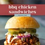 A BBQ chicken sandwich topped with coleslaw and pickle slices on a bun, served on a black plate.