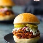 Pulled BBQ chicken sandwich topped with coleslaw and pickle slices on a black plate, with another sandwich blurred in the background.