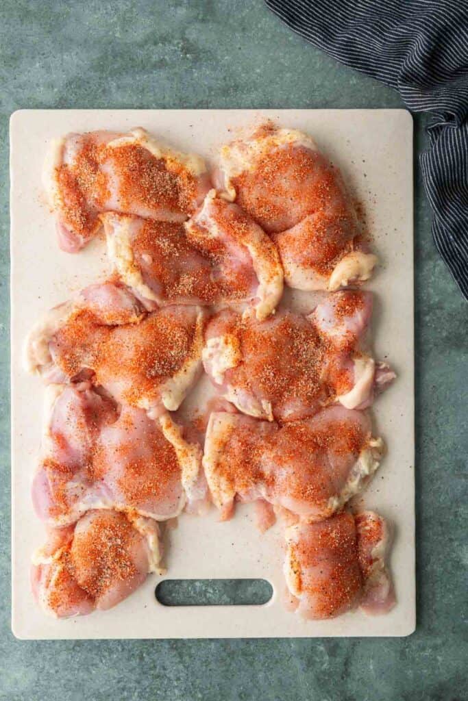 Raw chicken thighs on a white cutting board, sprinkled with a reddish-brown spice mix ready for a flavorful bbq chicken sandwich, on a green countertop with a striped cloth nearby.