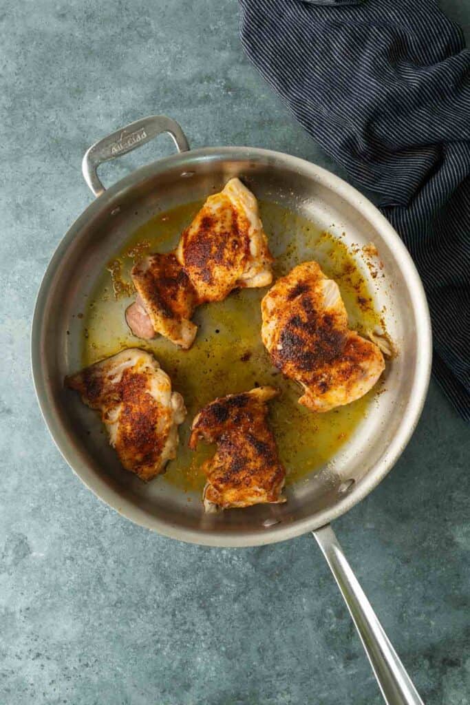 Four seasoned chicken thighs are searing in a stainless steel frying pan with oil, on a gray countertop next to a dark-striped cloth, ready to be shredded for the ultimate bbq chicken sandwich.