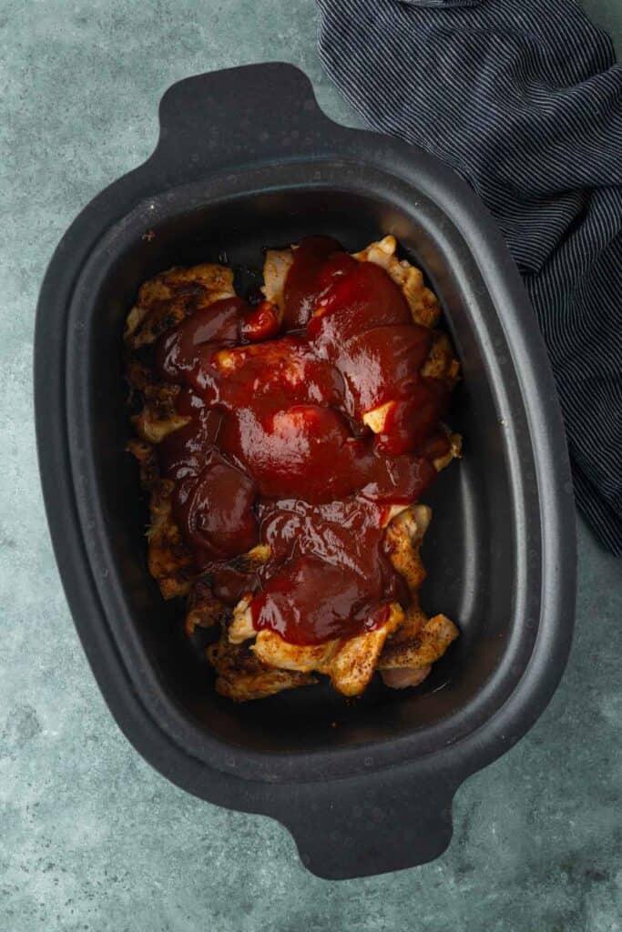Slow cooker filled with cooked chicken thighs topped with a thick layer of barbecue sauce, perfect for piling onto a bun to create a classic bbq chicken sandwich, placed on a textured gray surface next to a dark cloth napkin.