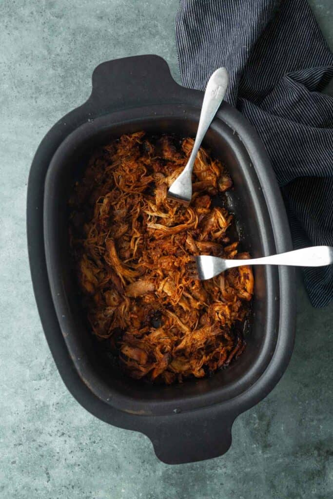 Shredded cooked meat in a black slow cooker with two forks, perfect for making a bbq chicken sandwich, placed on a gray surface next to a dark striped cloth.