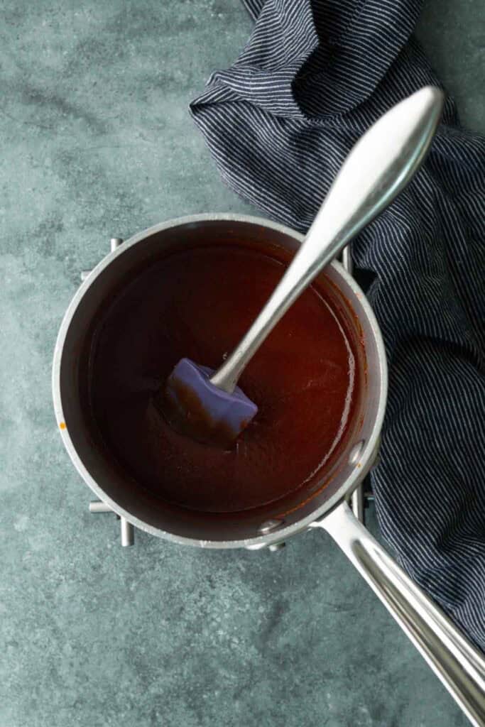 A saucepan filled with dark brown sauce, perfect for a bbq chicken sandwich, is stirred with a spatula and sits on a green countertop next to a striped cloth.