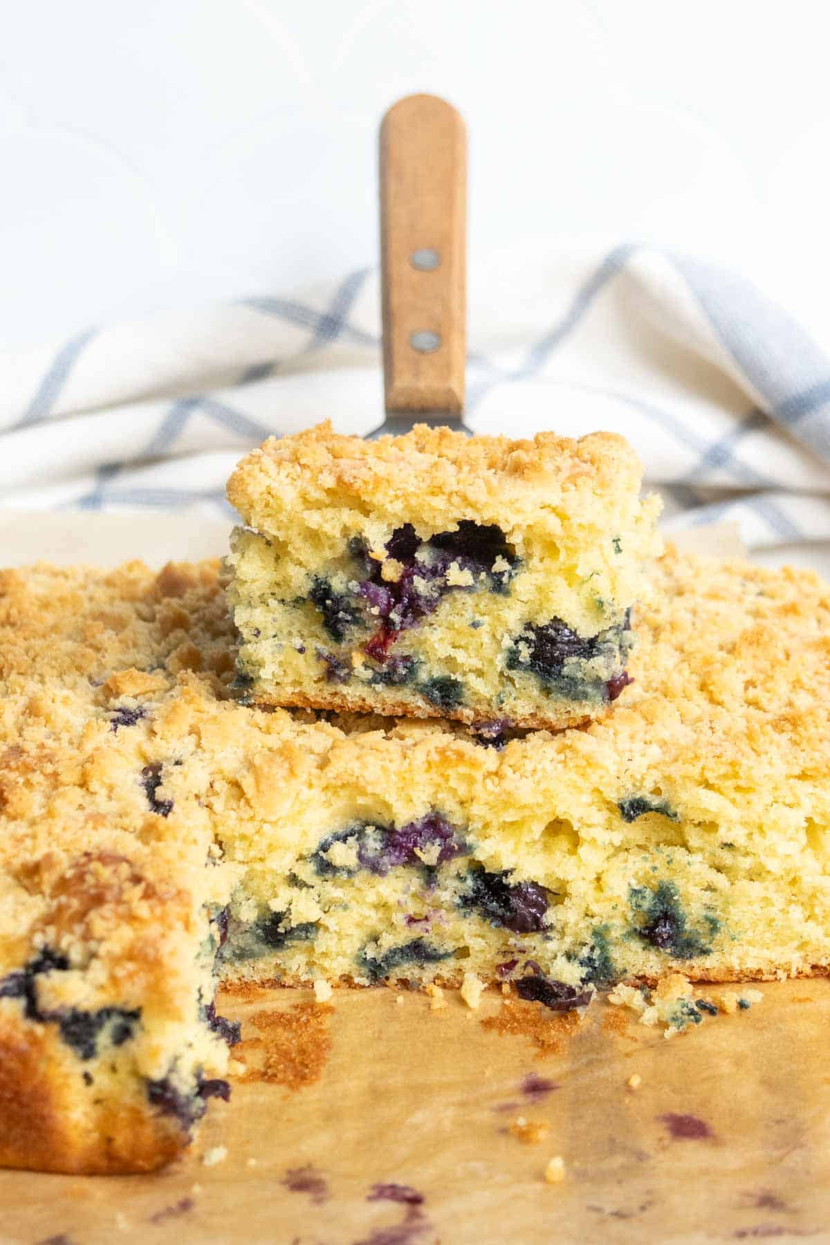 A slice of blueberry crumb cake is placed on top of the remaining cake, showing its crumbly topping and blueberry-filled interior. A knife stands upright in the background.