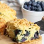A slice of blueberry crumb cake sits on a wooden board with fresh blueberries in a white bowl in the background.