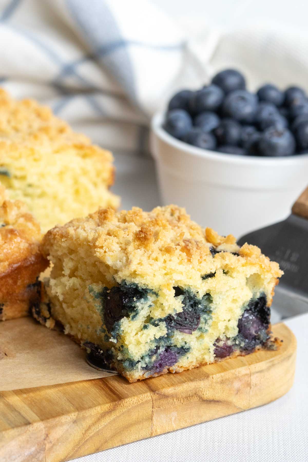 A slice of blueberry crumb cake sits on a wooden board with fresh blueberries in a white bowl in the background.