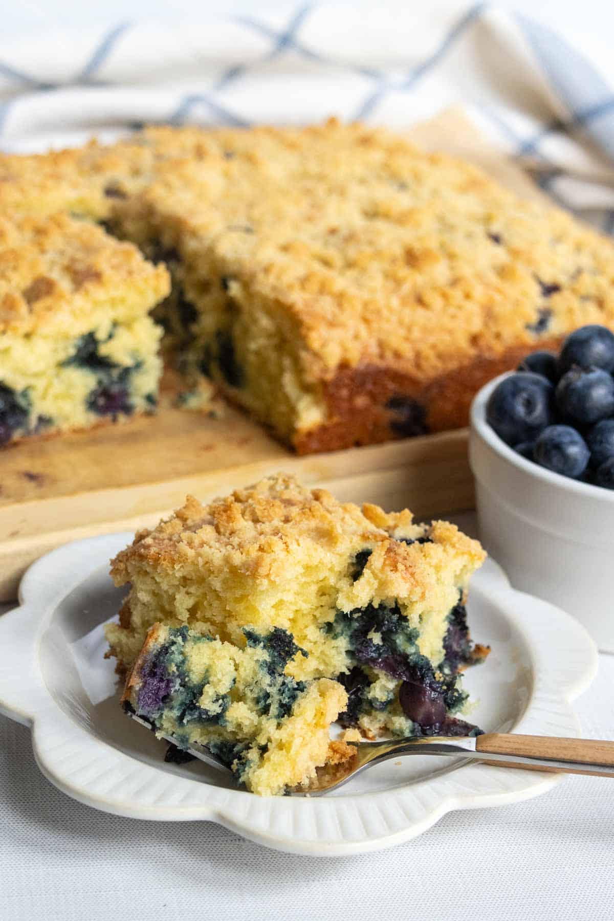 A slice of blueberry coffee cake sits on a white plate with a fork, next to a bowl of fresh blueberries and the remaining cake in the background.