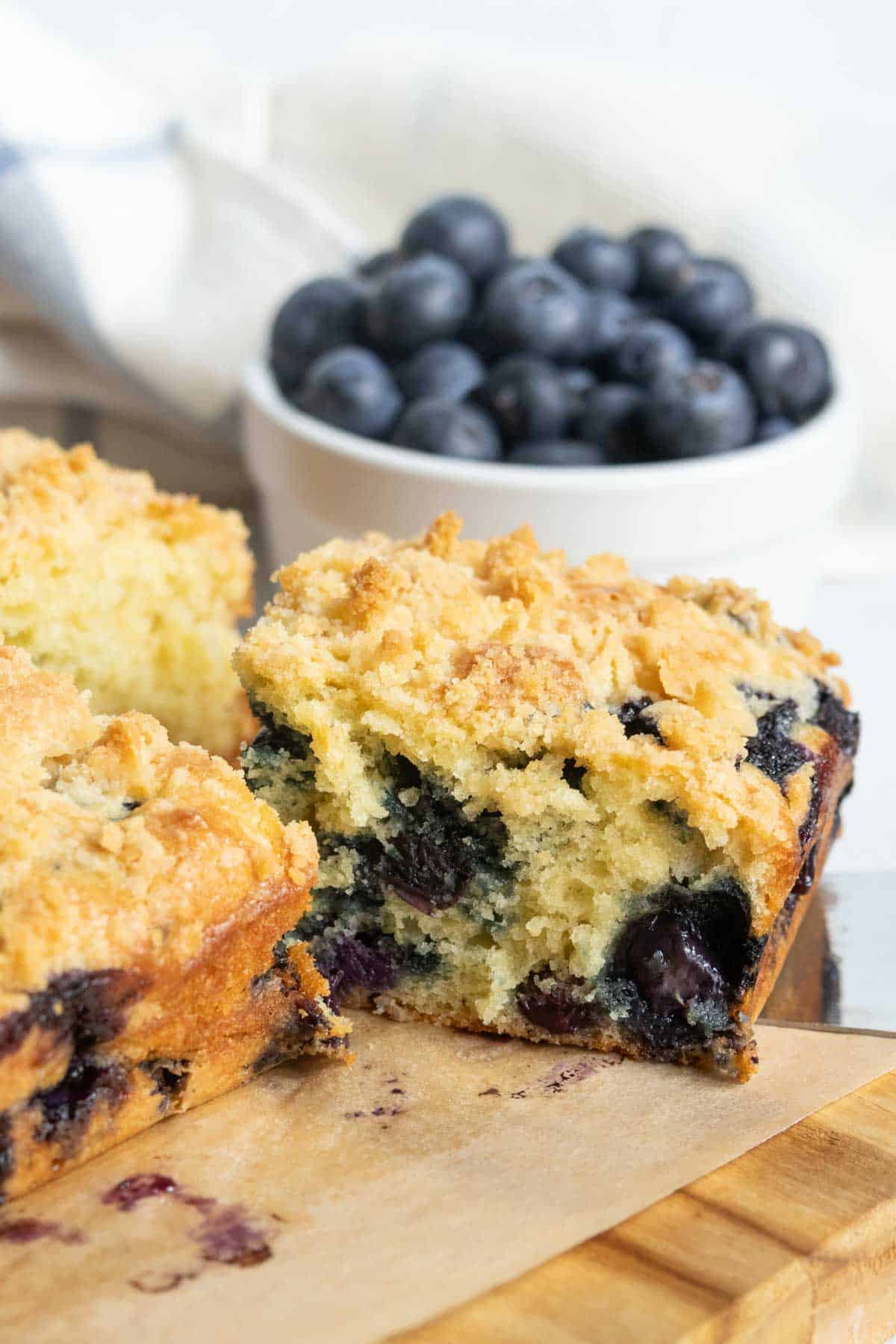A close-up of a sliced blueberry crumb cake on a wooden board, with a bowl of fresh blueberries in the background.