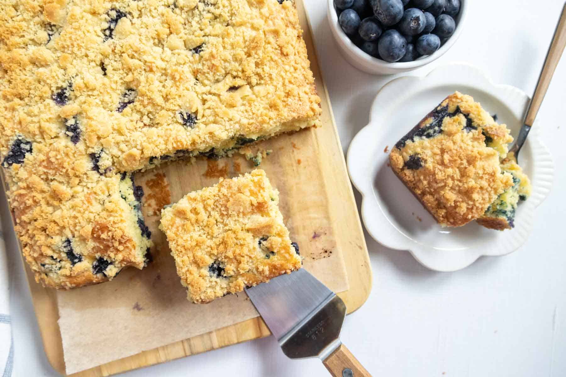 A slab of blueberry crumb cake on a wooden board, with one square piece being served, and a bowl of blueberries and a plated piece beside it.