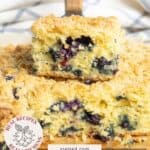 Two slices of blueberry coffee cake stacked on a plate, with a crumbly topping and visible blueberries, in front of a white towel.
