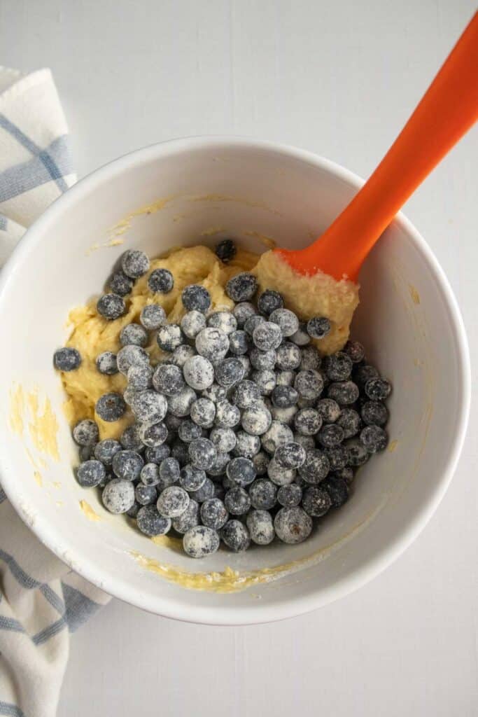 A white bowl containing batter and flour-dusted blueberries being stirred with an orange spatula.