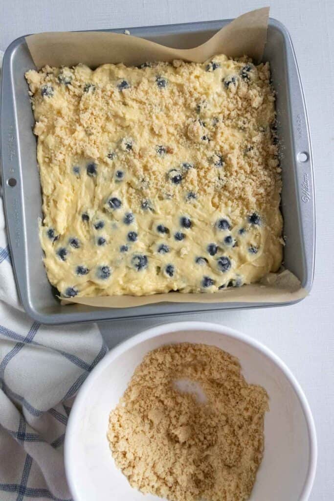 Blueberry coffee cake batter in a pan with crumble topping half on and half in a bowl.