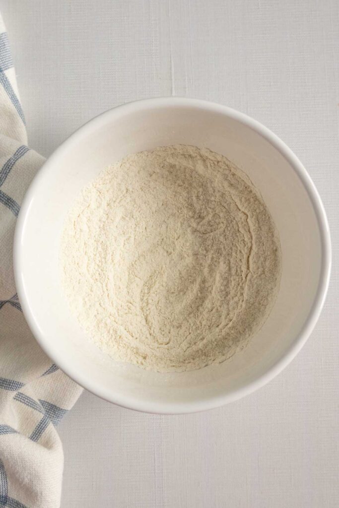 A white bowl containing flour with a kitchen towel next to it.