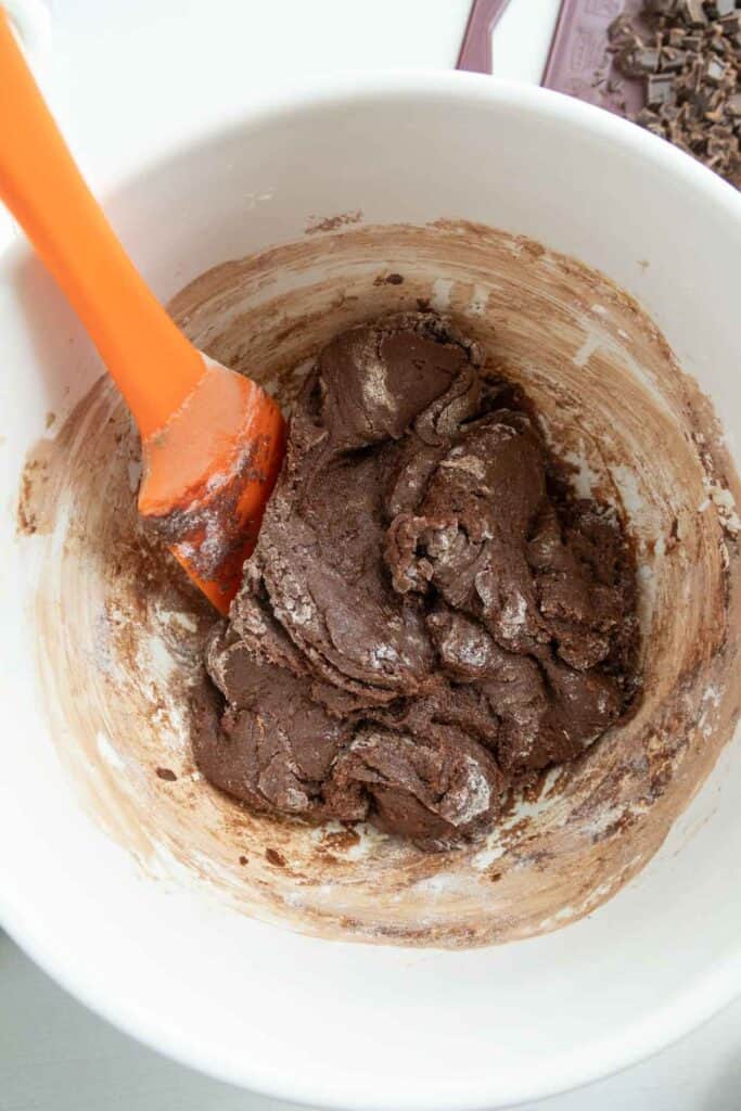 An orange spatula stirring chocolate cookie dough in a white bowl.