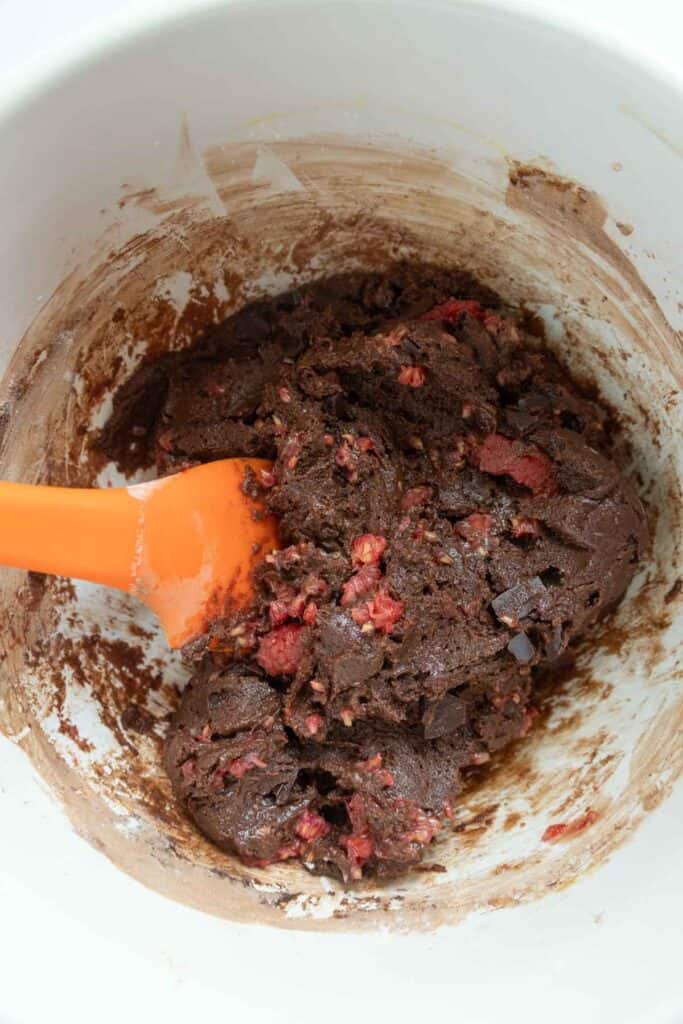An orange spatula stirring chocolate raspberry cookie dough in a white bowl.