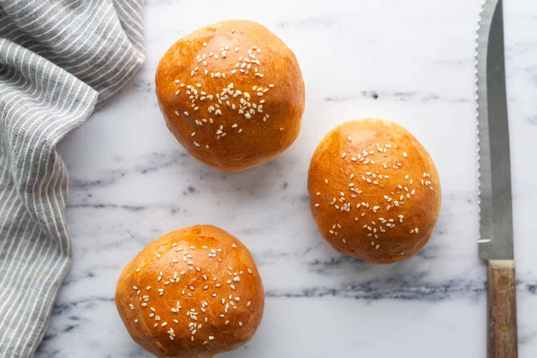 Three burger buns with sesame seed tops next to a knife.