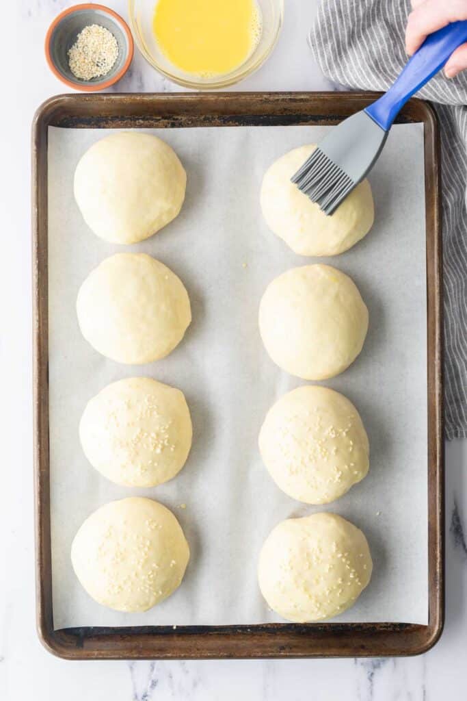 A hand brushing egg wash onto unbaked burger buns on a sheet pan.
