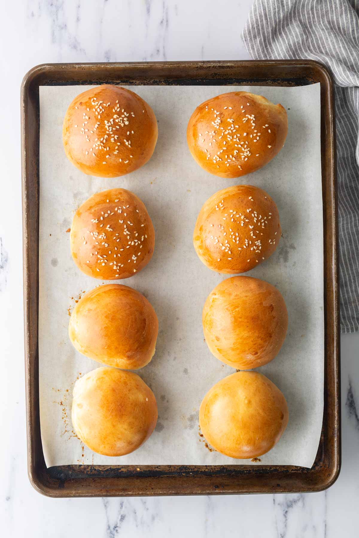 Baked burger buns on a sheet pan, with half the buns sprinkled with sesame seeds.