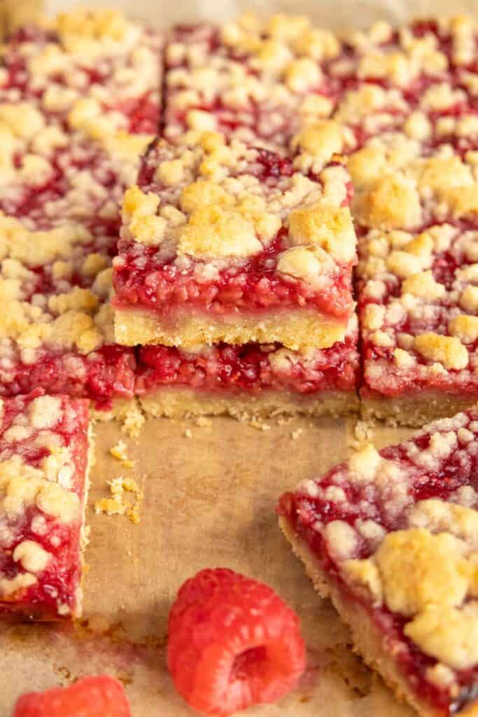 Raspberry Bars Raspberry crumble bars are stacked on parchment paper, with a few fresh raspberries in front and a piece of the delicious raspberry bars pulled slightly out from the batch.