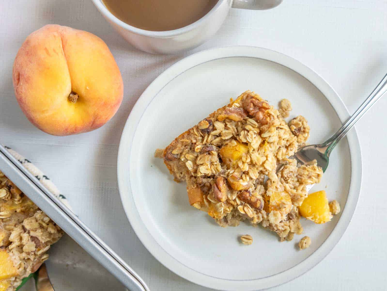 A plate with a serving of baked oatmeal and a fork, next to a whole peach, a baking dish, and a cup of coffee on a white table.