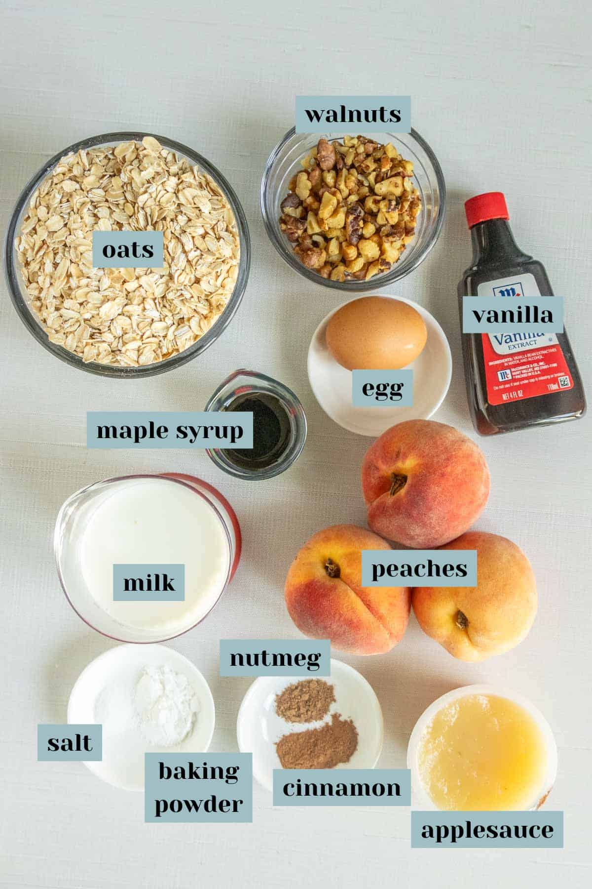 Top-down view of labeled breakfast ingredients including oats, walnuts, maple syrup, milk, egg, vanilla, peaches, nutmeg, cinnamon, salt, baking powder, and applesauce on a white surface.