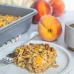 A slice of baked oatmeal with peaches and oats on a plate with a fork, fresh peaches, a baking dish, and a cup in the background.
