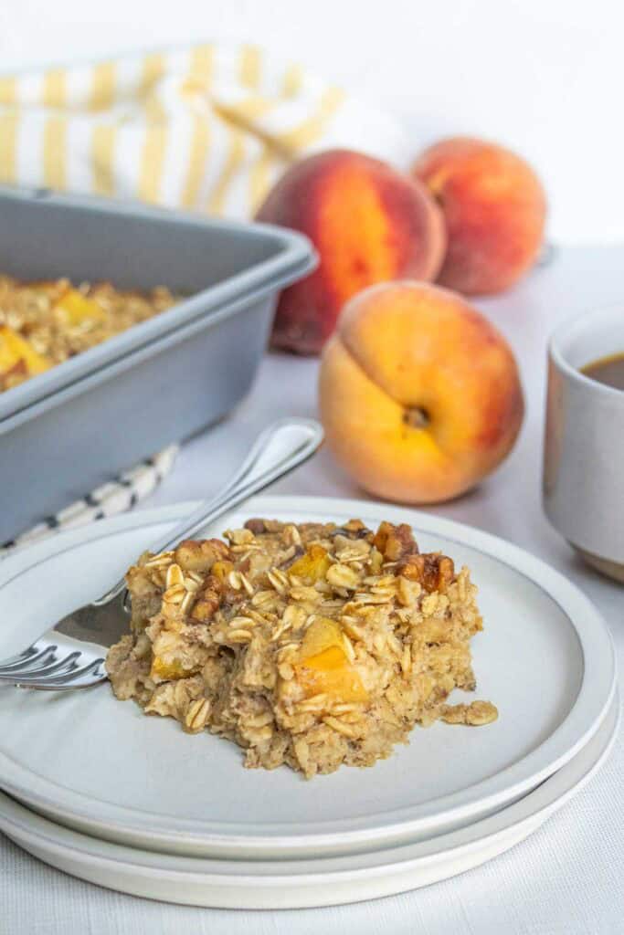 Baked Peach Oatmeal A slice of baked oatmeal with peaches and oats on a plate with a fork, fresh peaches, a baking dish, and a cup in the background.