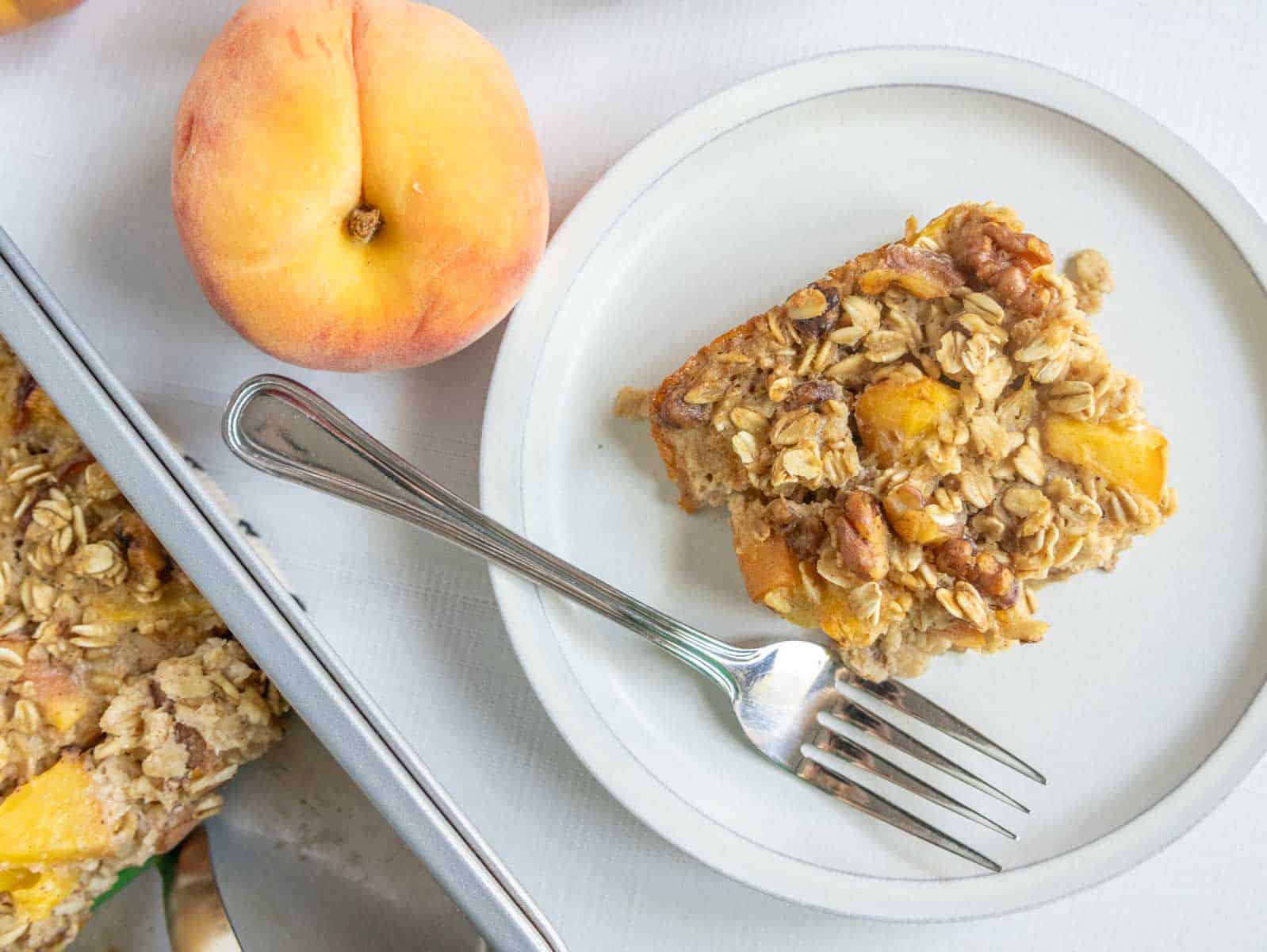 A slice of baked oatmeal with peach pieces on a white plate with a fork, next to a whole peach and a baking tray.