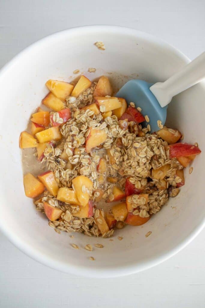 A white bowl contains chopped peaches, rolled oats, and liquid being mixed with a blue silicone spatula.