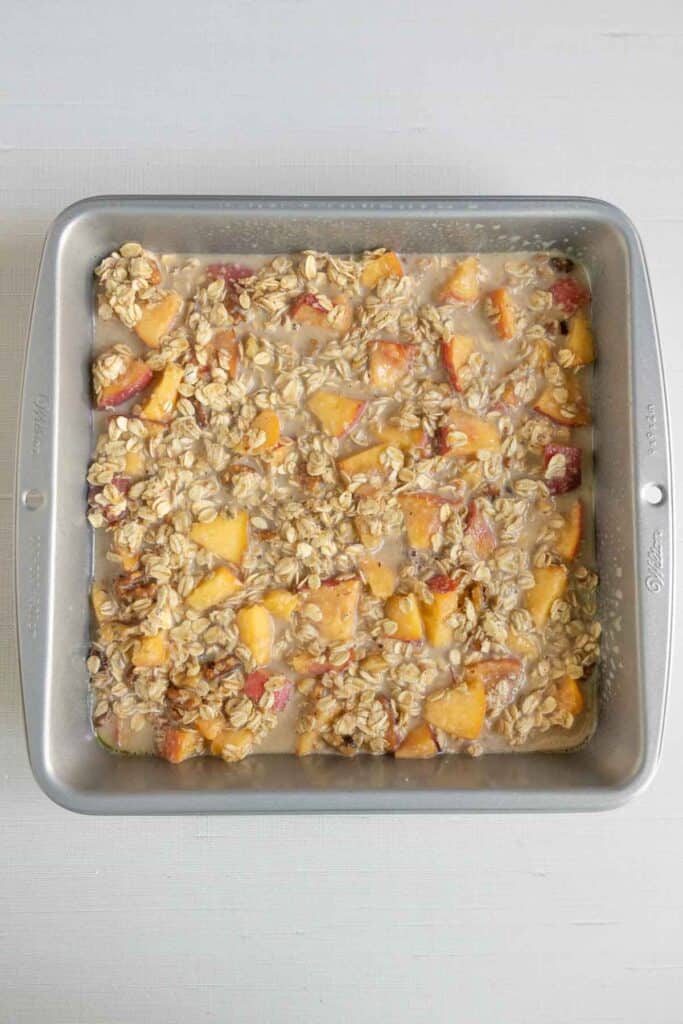 A square metal baking pan filled with an unbaked mixture of oats, sliced peaches, and liquid, ready to be baked.