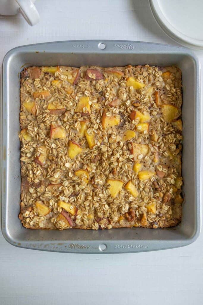 A square baking pan filled with baked oatmeal topped with chunks of peaches sits on a white surface. Plates are partially visible in the upper right corner.