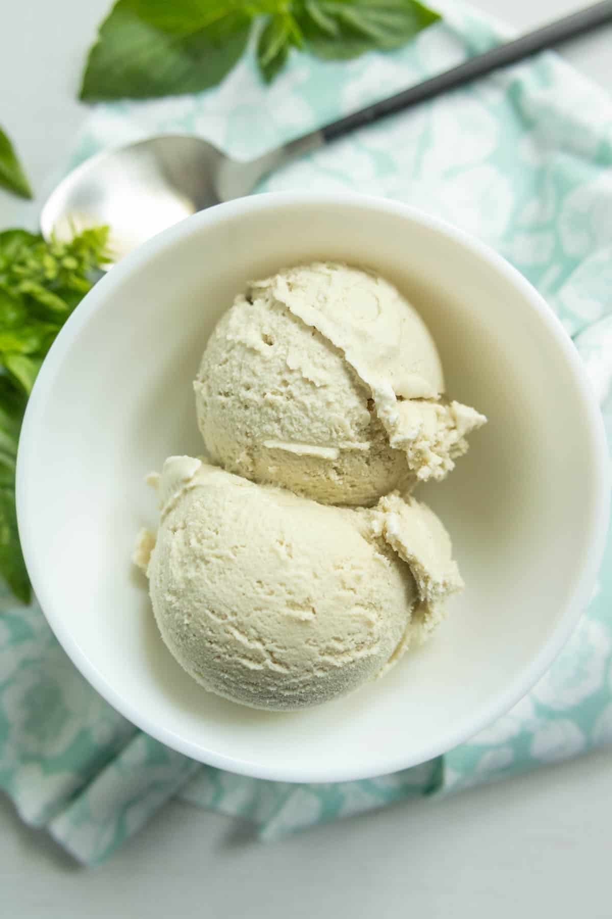 Two scoops of vanilla ice cream in a white bowl, placed on a light patterned cloth with a spoon and green leaves nearby.