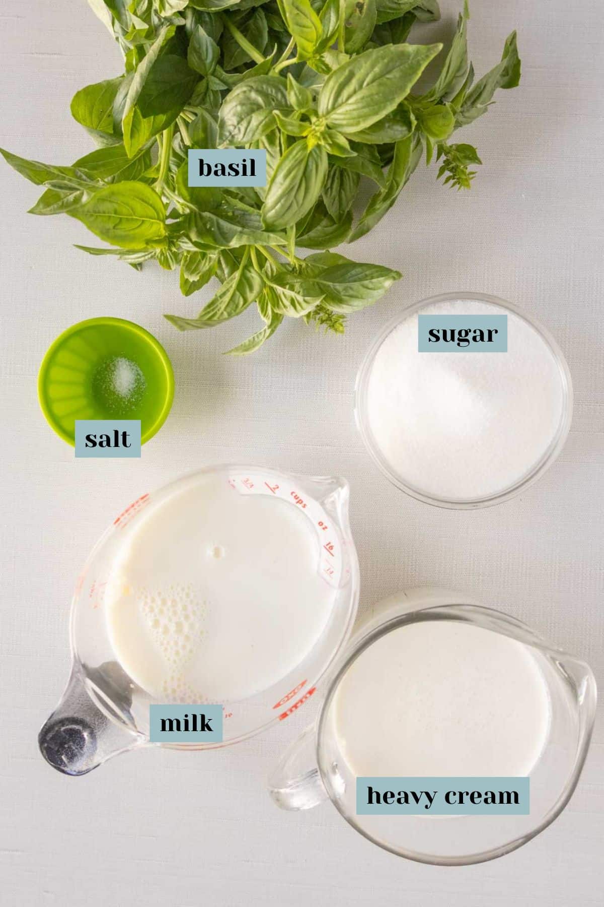Overhead view of basil, a cup of sugar, a cup of heavy cream, a cup of milk, and a small container of salt on a white surface, each labeled.