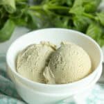 Two scoops of light-colored ice cream in a white bowl, placed on a light blue and white patterned cloth, with green leaves in the background.