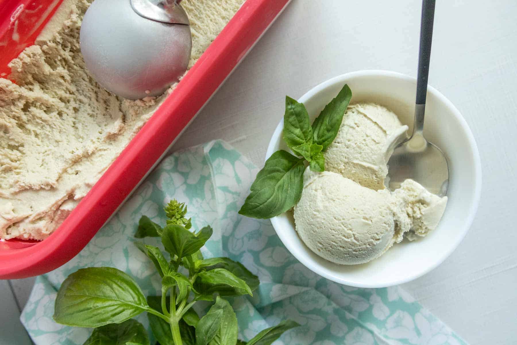Bowl of green ice cream with a spoon and basil leaves, next to a red container of ice cream and fresh basil on a patterned cloth.