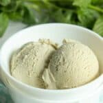 Two scoops of pale green basil ice cream in a white bowl, with fresh basil leaves in the background.