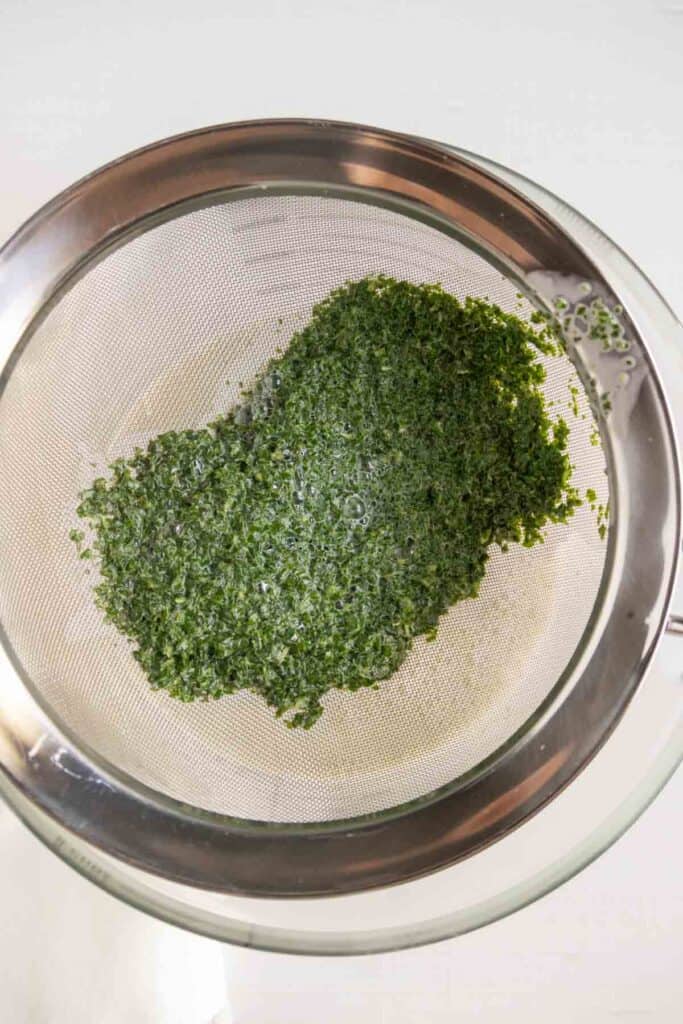 Finely chopped green herbs are draining in a fine mesh strainer placed over a glass bowl.