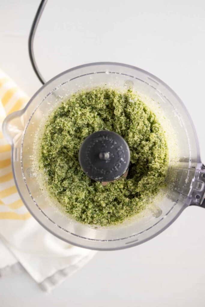 A top-down view of a food processor containing a coarse green mixture on a white surface next to a striped towel.